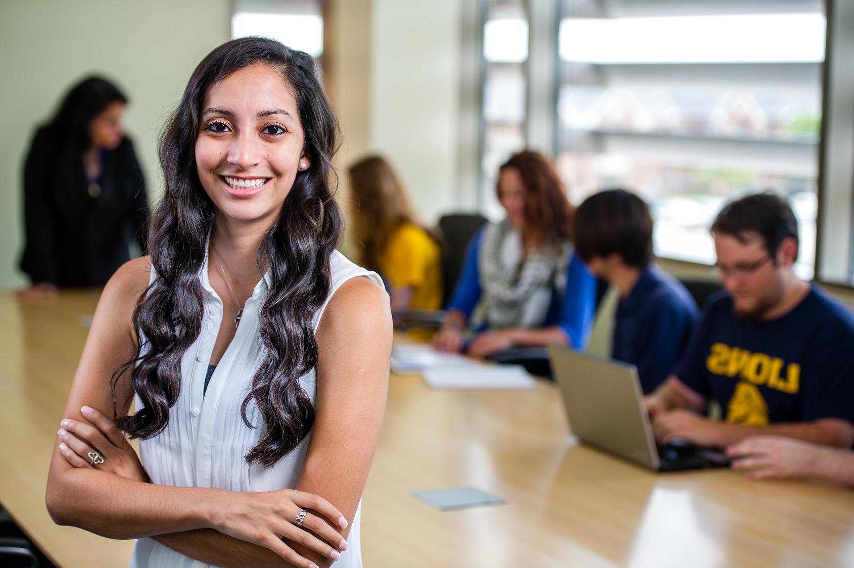 Student smiling posing for photo in front of a desk where other students are sitting down working together.